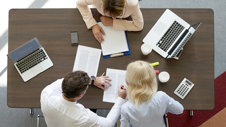 Overhead view of people reviewing documents and working together at a table with laptops, notebooks, and coffee cups.