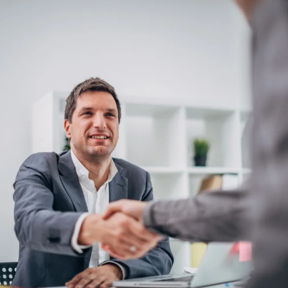 A person in professional attire sitting at a desk and shaking hands with another individual across the table in a modern office setting.